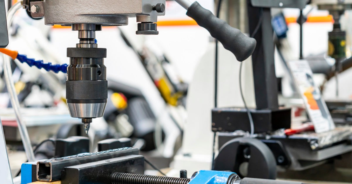 A close-up view of a vertical drill press machine in a workshop. The drill is hovering over a piece of metal.