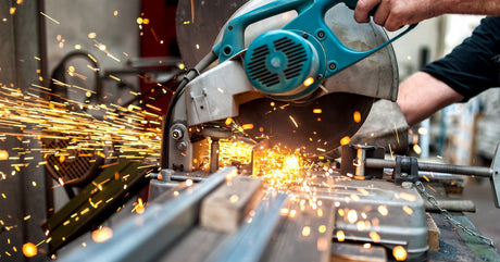 A worker using a metal cutting circular saw to cut through multiple thick metal poles in a work shop.