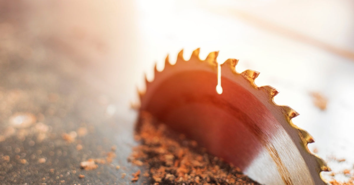 A close-up view of a stainless steel circular saw blade cutting a piece of wood. The edge of the blade has large teeth.