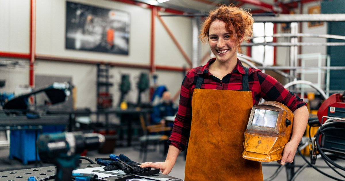 A woman is standing in a workshop and smiling at the camera. She is wearing protective equipment and holding a helmet.