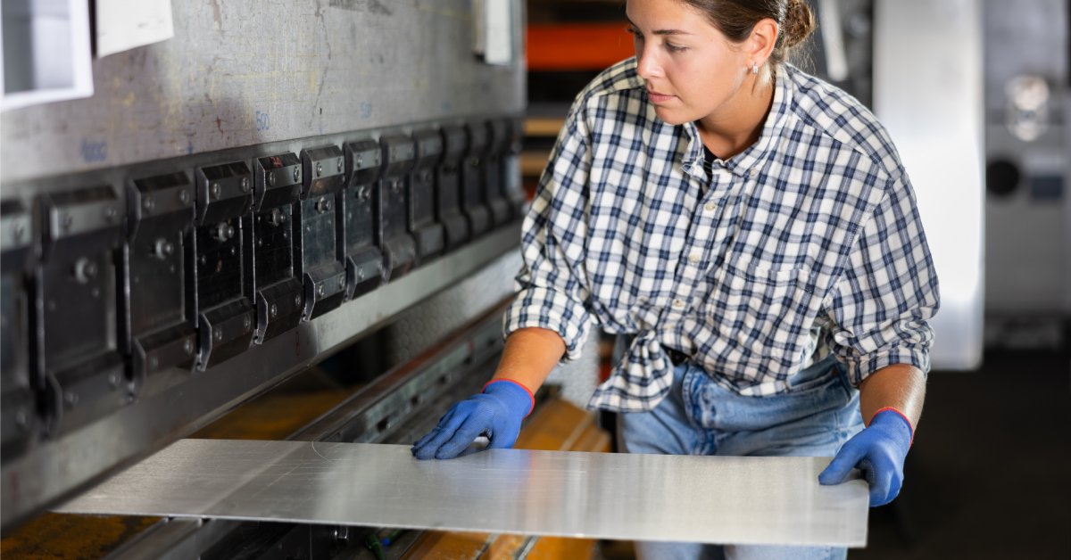 A woman in a plaid shirt wearing blue gloves while feeding a flat sheet of metal through a large press brake.