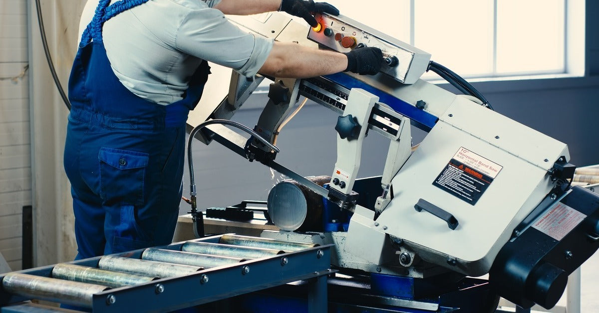 A man wearing blue overalls and black gloves while using a metal band saw to cut a piece of cylindrical metal.