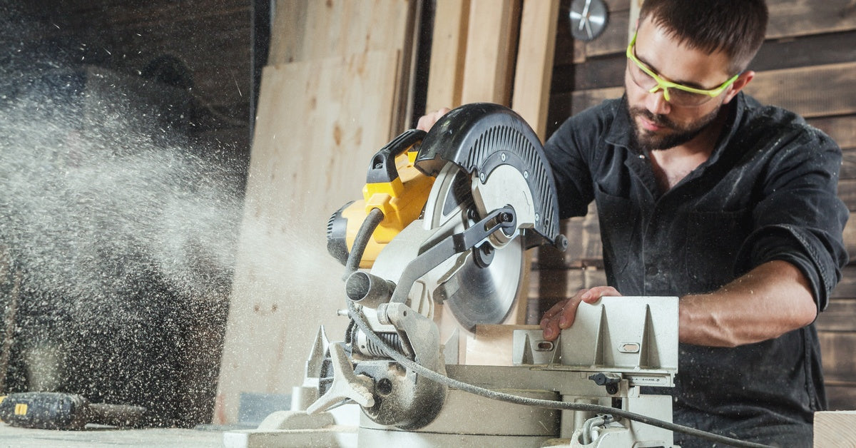 A man wearing yellow protective glasses uses a circular saw to cut through multiple planks of wood.