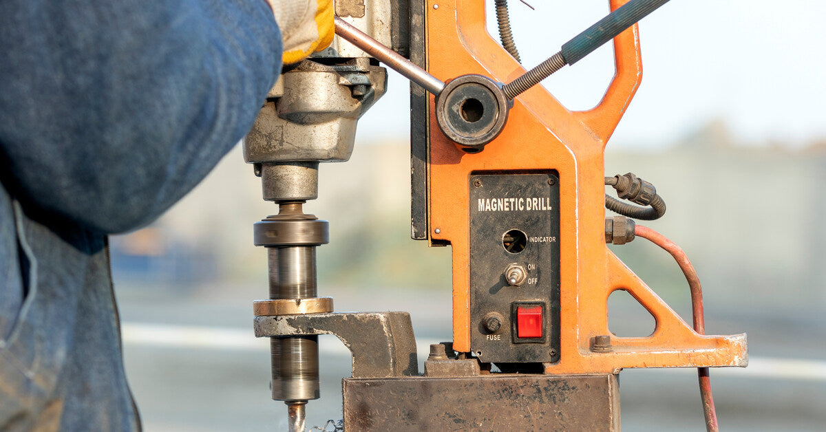 A worker using an orange-colored vertical drilling machine to drill into a piece of steel at a construction site.