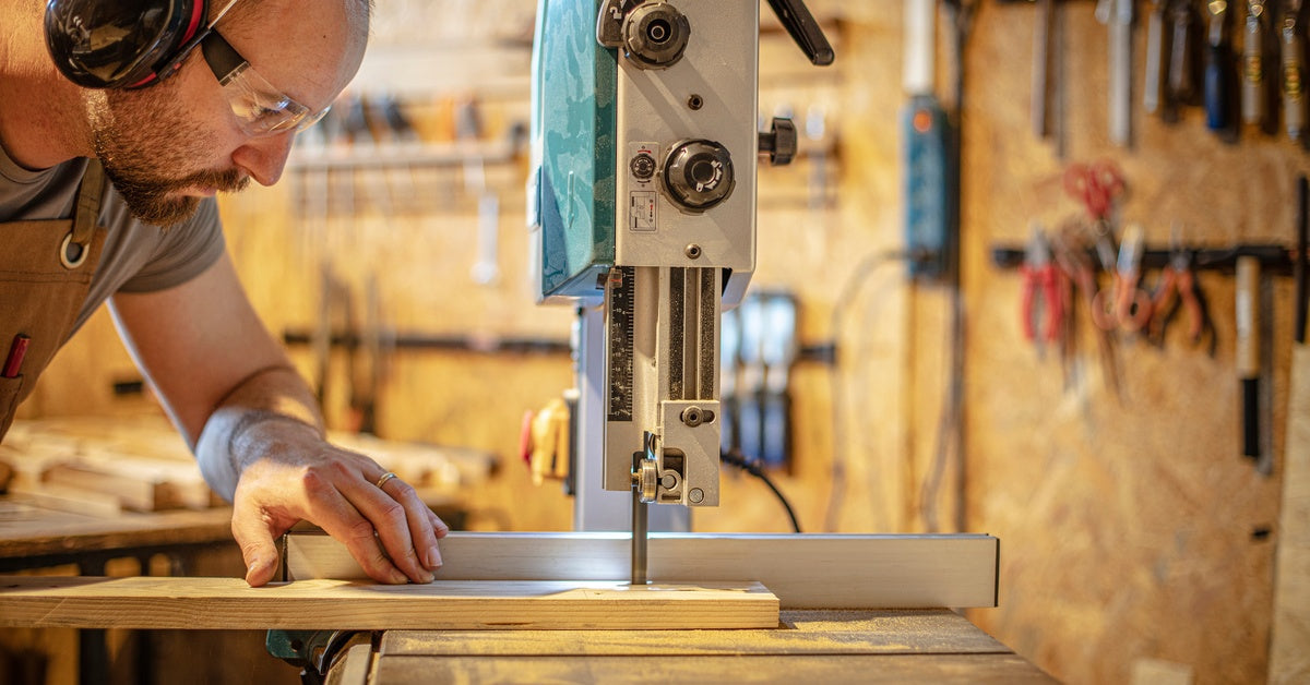 A carpenter wearing protective glasses while using a band saw in a workshop to cut through a thin plank of wood.