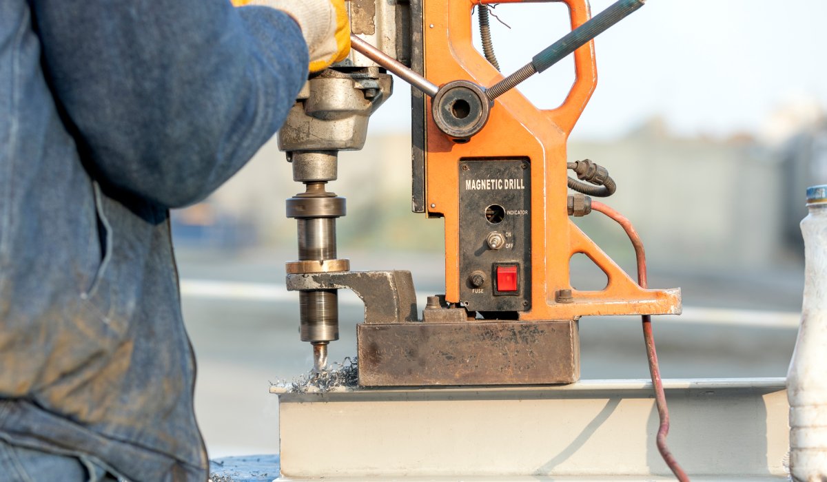 A worker wearing a blue jumpsuit uses an industrial vertical drilling machine to drill through a metal object.