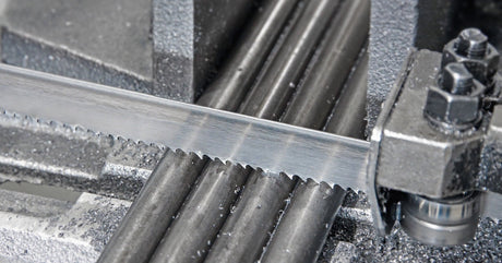 A close-up of an industrial automatic band saw cutting through four pipes surrounded by metal shards in a workshop.