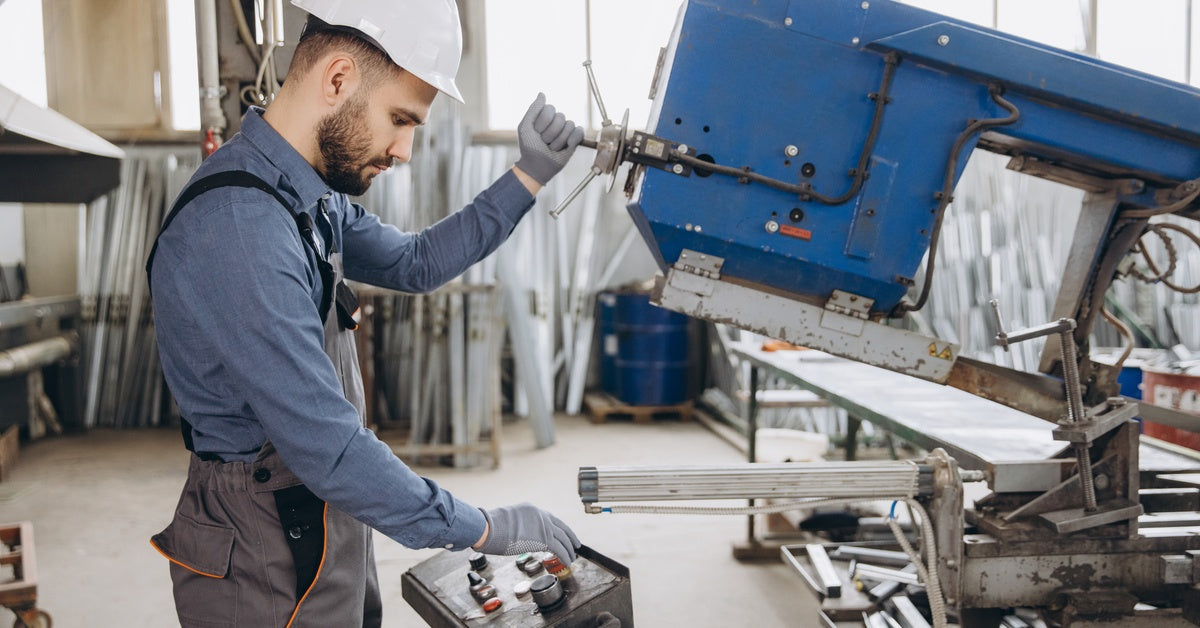 A worker in overalls and a protective white hard hat standing while operating a metal band saw in a workshop.