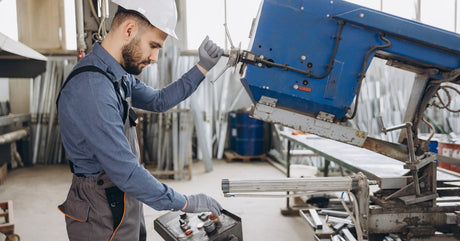 A worker in overalls and a protective white hard hat standing while operating a metal band saw in a workshop.