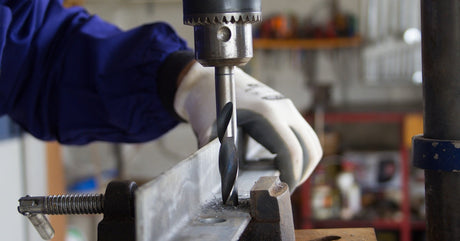A worker wearing white gloves operates an industrial drilling machine to drill holes through a metal bar.