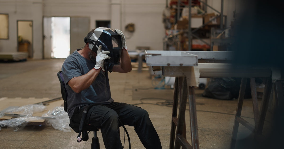 A welder sitting in a metalworking shop. They are wearing white gloves and removing a welding helmet from their head.