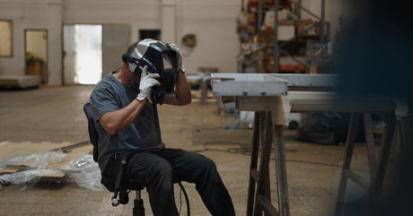 A welder sitting in a metalworking shop. They are wearing white gloves and removing a welding helmet from their head.