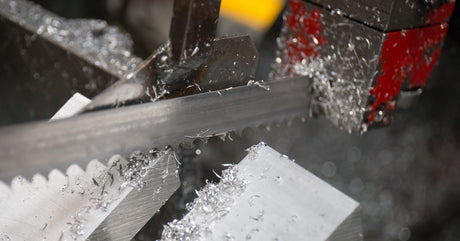 A bandsaw machine with a metal blade cuts through a piece of aluminum. Metal shavings fly in the air around the blade.