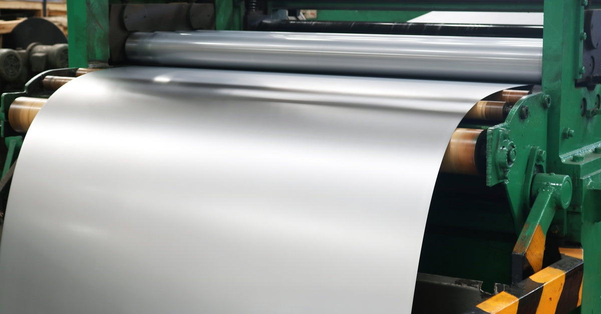A shiny sheet of metal comes out of a cold rolling machine. The machine is green with black and yellow guard rails.