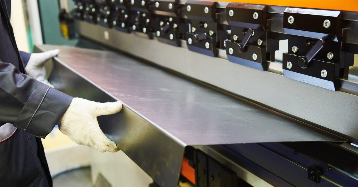 A worker wearing gloves guides sheet metal into an orange industrial press brake in a bright workshop.